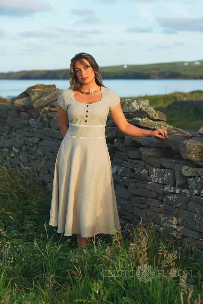 Square neck beige midi dress with pockets, styled as a natural fibre folklore dress against a coastal stone wall