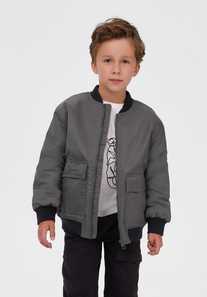 A young boy wearing a gray bomber jacket with black ribbed trim over a white graphic t-shirt and black pants, standing against a light gray studio background.