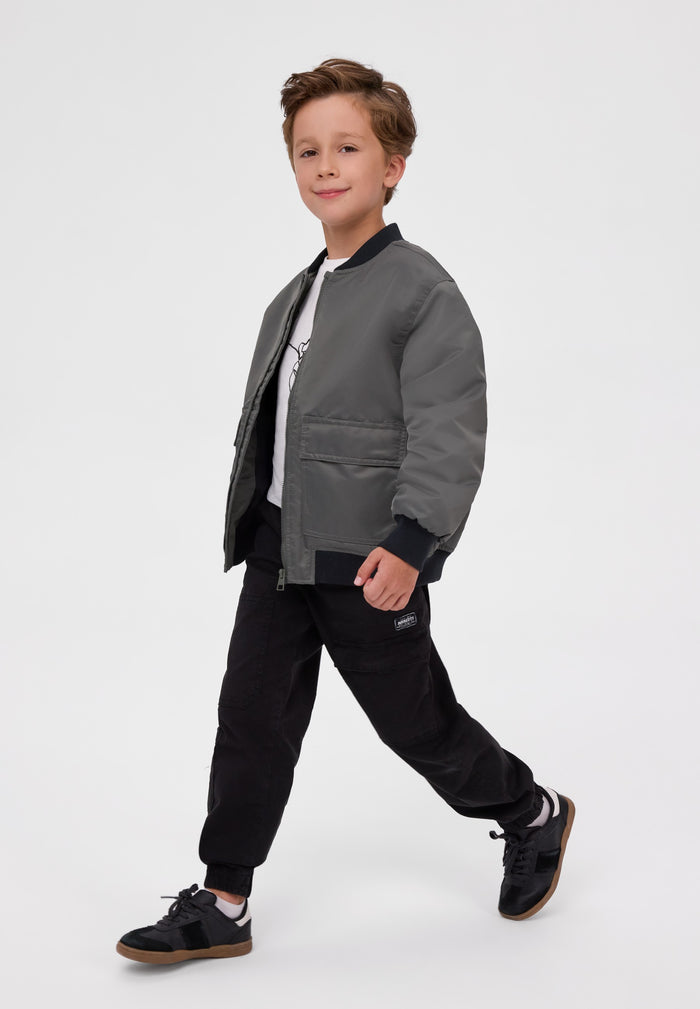 A young boy modeling a dark gray bomber jacket, styled with black joggers, a white t-shirt, and black sneakers, set against a plain light gray studio background.
