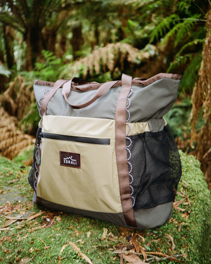 A large tote bag with a beige and gray body, brown straps, and a black mesh side pocket, resting on a mossy log in a lush green forest.