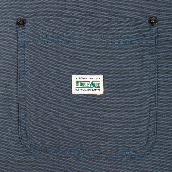 Close-up of a blue canvas work shirt pocket with two metal snap buttons and a white Dubbleware brand label stitched in the center.