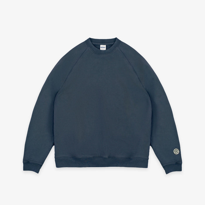 A dark slate blue crewneck sweatshirt with raglan sleeves, shown flat against a light gray background. A small circular logo is printed on the left sleeve cuff.