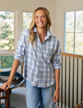 Woman wearing a blue plaid shirt and light blue pants standing in a room with large windows.