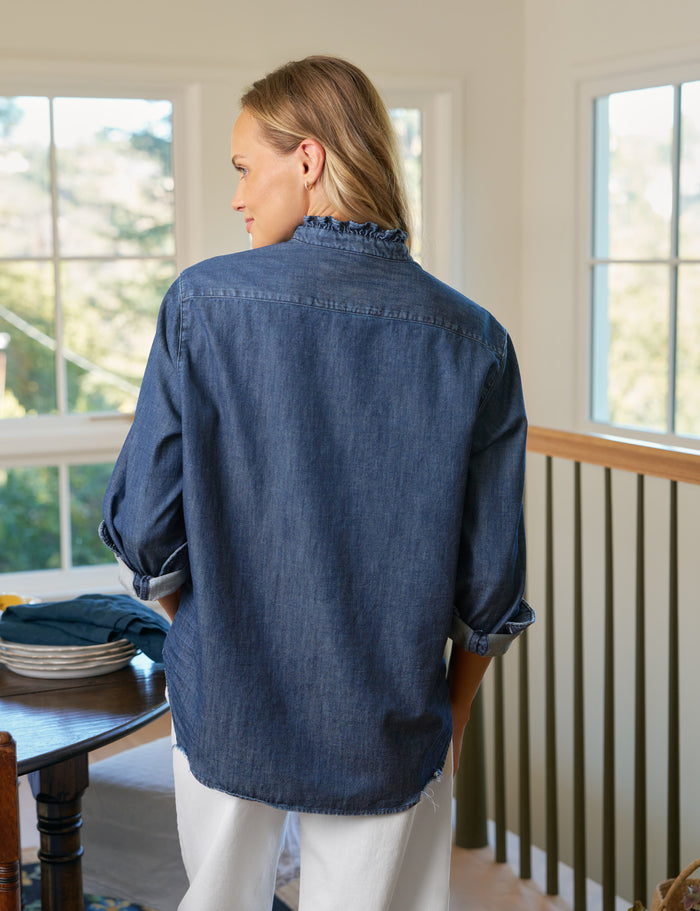 Person wearing a blue denim shirt standing in a room with large windows.
