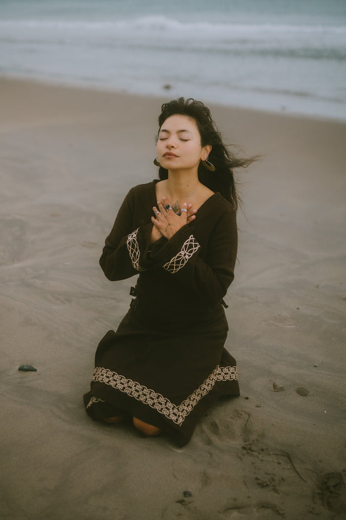 Folklore-inspired brown long sleeve wrap top with Celtic knotwork cuffs, worn with a matching skirt by the sea.