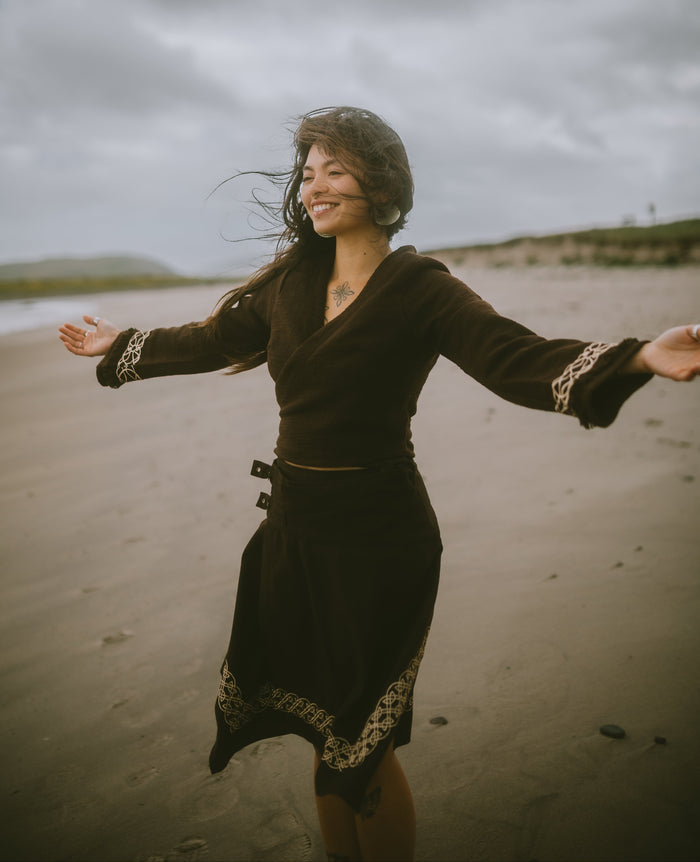 Brown folklore crop top with adjustable wrap fit and Celtic embroidered cuffs designed by Celtic Fusion and styled on a moody coastal beach with windswept hair