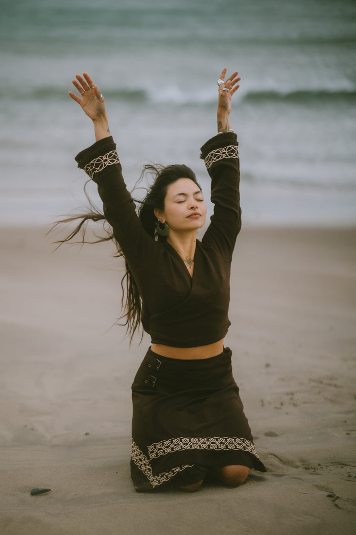 Elva Brown Celtic Wrap Cardigan styled with a matching embroidered skirt on a windswept Irish beach.