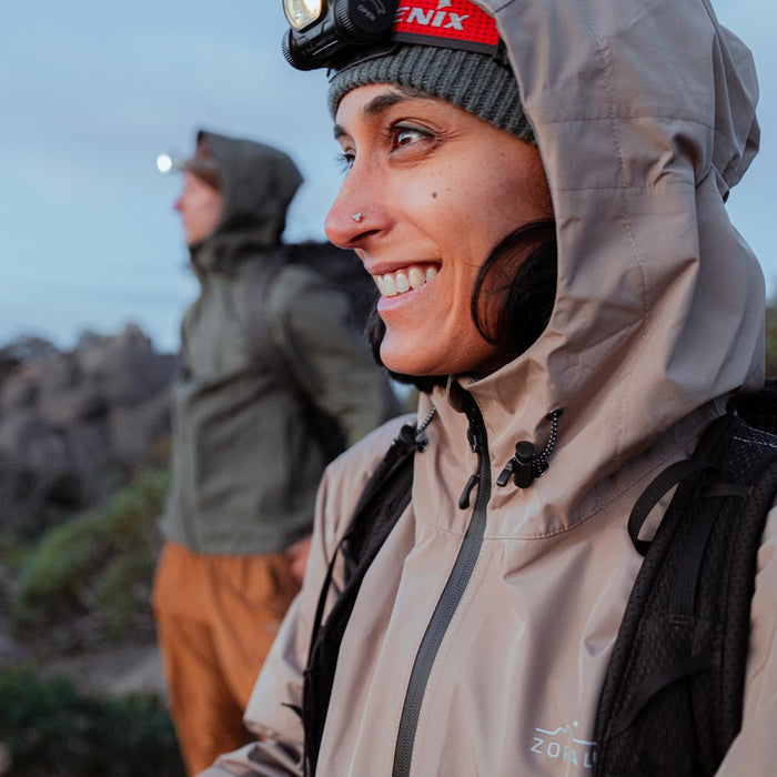Person wearing a beige jacket with a hood, smiling outdoors.
