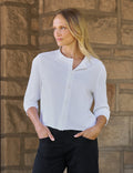 A woman wearing a white long-sleeve button-up shirt and black pants, standing against a rustic stone wall.