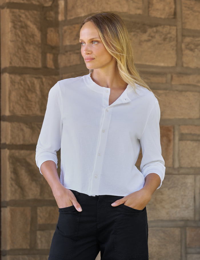 A woman wearing a white long-sleeve button-up shirt and black pants, standing against a rustic stone wall.