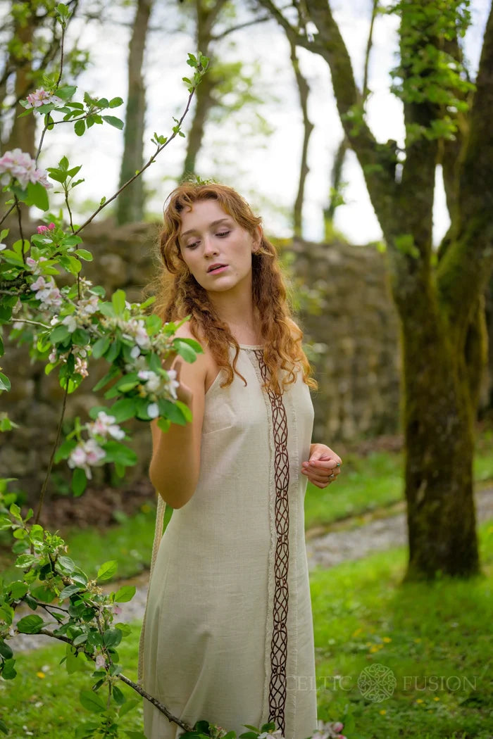 Sleeveless white boho maxi dress with Celtic knotwork embroidery panel, photographed in a blossom garden