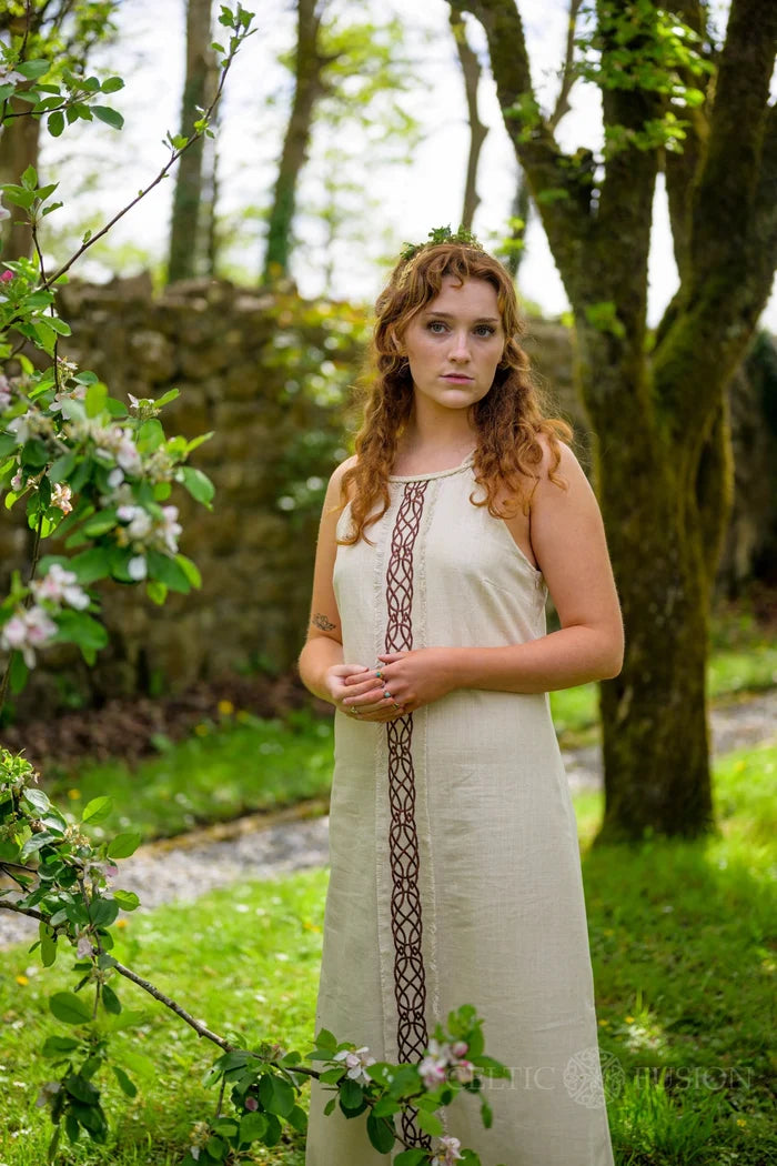 Natural folklore maxi dress with rope tie straps and vertical Celtic embroidered panel, styled in an orchard setting.