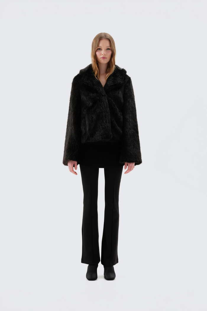 Black faux fur jacket with notch lapel and cropped fit, worn open over a black top and flared trousers, front view