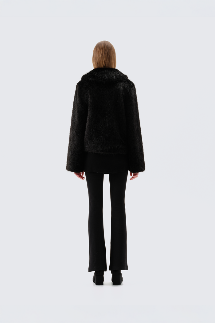 Black faux fur jacket with notch lapel and cropped fit, worn open over a black top and flared trousers, back view