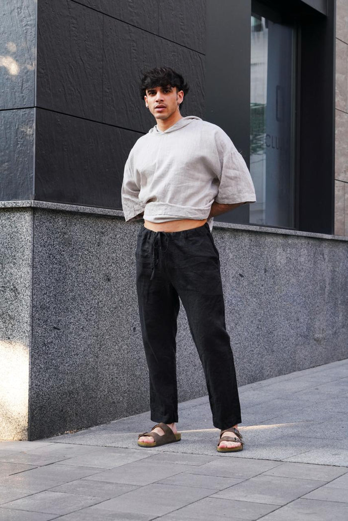 A man is wearing a beige linen hoodie and black linen pants standing against a dark building.