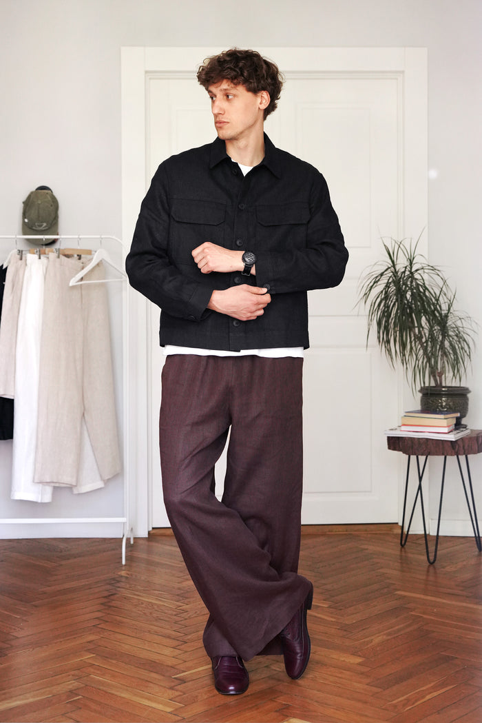 Man wearing a black jacket and brown pants standing in a room with wooden flooring and white walls.