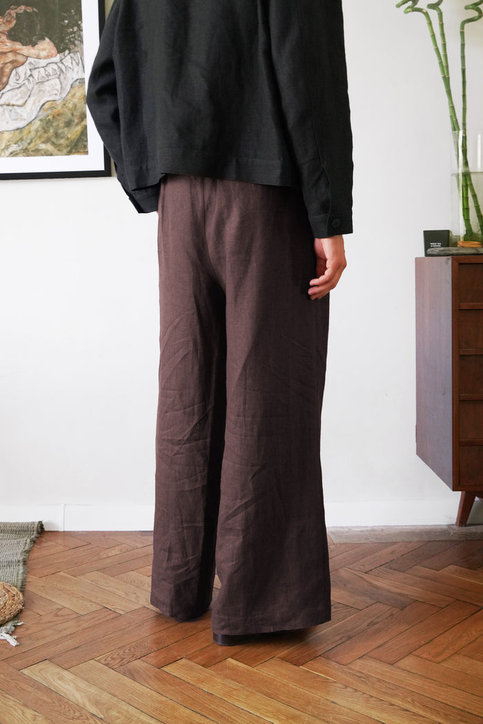 Person wearing brown wide-leg pants and a black top in a room with wooden flooring and a white wall.