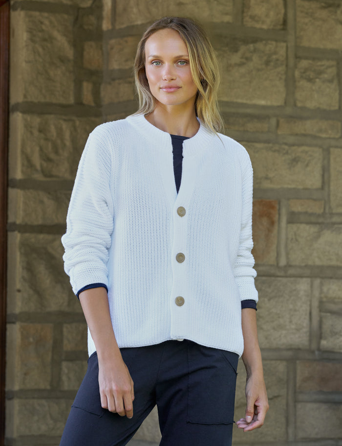 A woman wearing a white ribbed knit cardigan, paired with dark pants, standing in front of a stone wall.