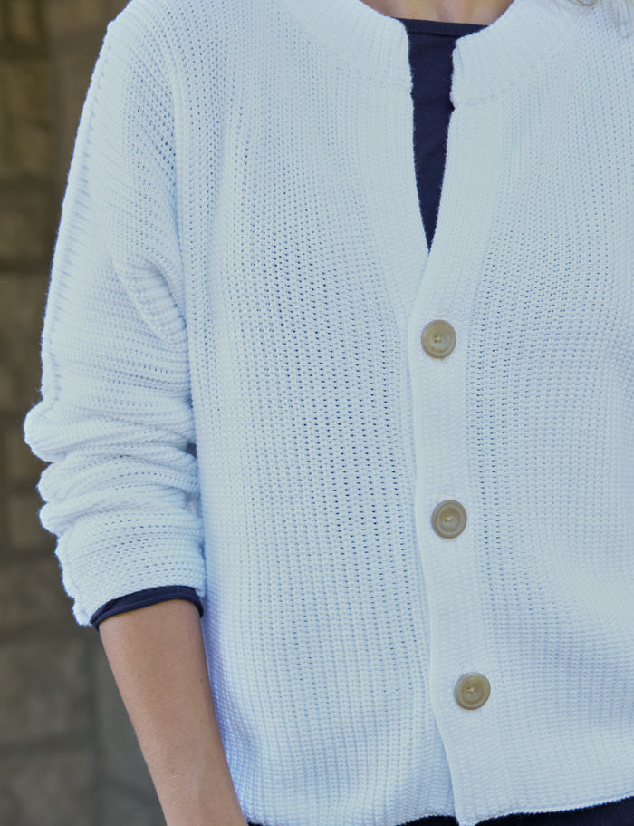 A close-up of a person wearing a white cardigan with three buttons over a dark navy top. The background is a softly blurred stone wall.