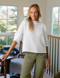Woman wearing a white sweater and green pants standing in a room with large windows.
