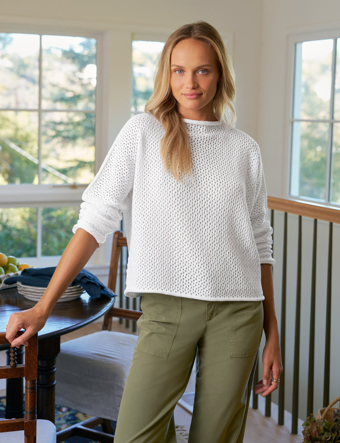 Woman wearing a white sweater and green pants standing in a room with large windows.