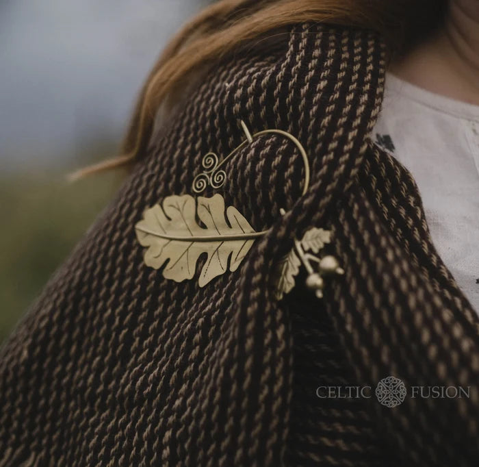 Close-up of brass oak leaf brooch fastening a handwoven wool blend shawl with fringe, showing textured woven detail