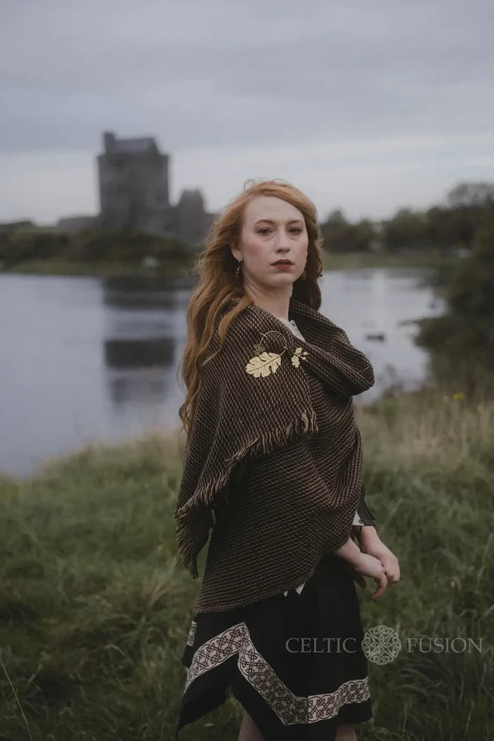 Brown wool blend shawl with fringe edge and brass leaf brooch, worn as a Celtic wrap in an outdoor heritage setting designed by Celtic Fusion